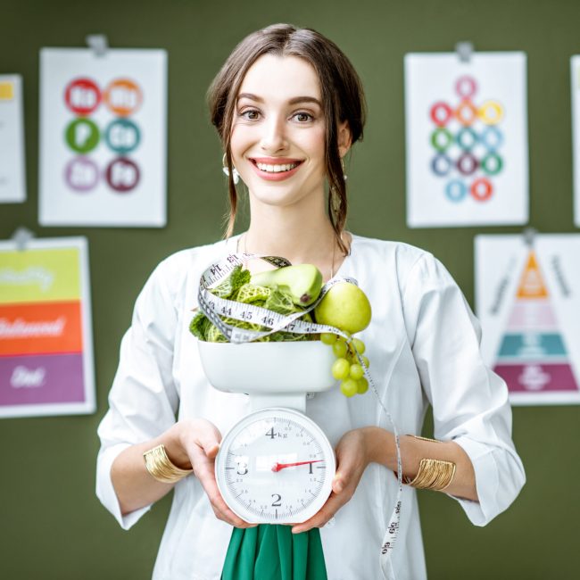 Young woman nutritionist in medical gown holding weights full of healthy products standing on the wall background with diet schemes